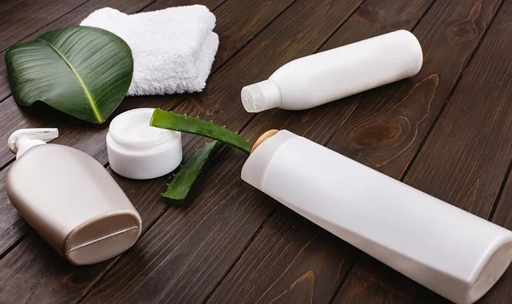 A variety of skincare bottles and a towel on a wooden surface with a green aloe leaf.
